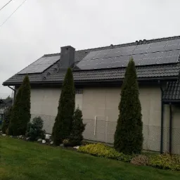 Dom jednorodzinny z ciemną dachówką i panelami słonecznymi na dachu, widok z ogrodu z tujami i trawnikiem.