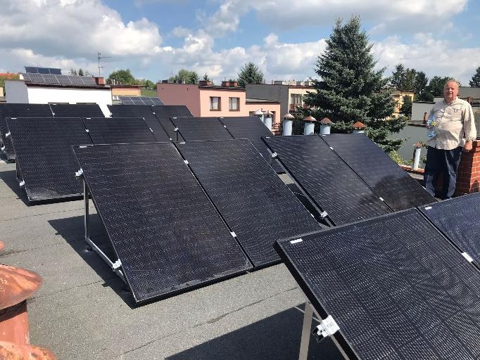 Instalacja paneli słonecznych na płaskim dachu budynku, z widoczną osobą stojącą obok komina, w tle budynki mieszkalne i drzewo