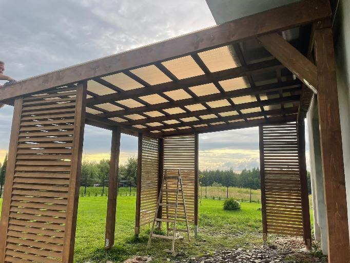 Drewniana pergola z ażurowymi ścianami i przezroczystym zadaszeniem w trakcie montażu, widoczna drabina i osoba pracująca na dachu, otoczenie zielone.
