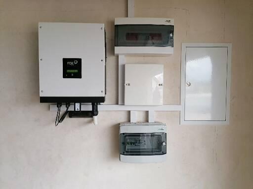 Biały falownik z czarnym panelem sterowania marki FOX, zamontowany na ścianie obok kilku skrzynek elektrycznych, połączonych białymi listwami.