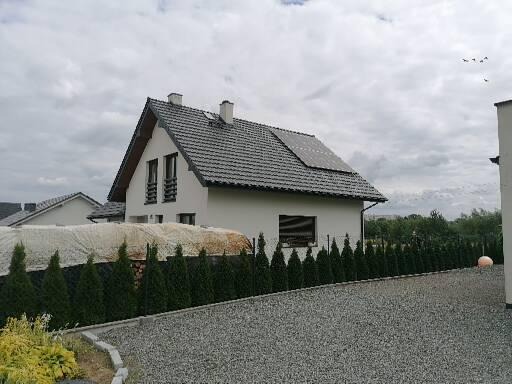 Dom jednorodzinny z szarą dachówką i panelami słonecznymi na dachu, widok z poziomu gruntu na zadbany ogród z tujami i szarą nawierzchnią z kamyczków.