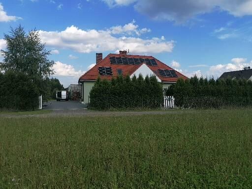 Dom jednorodzinny z czerwoną dachówką i panelami słonecznymi na dachu, widok zza trawnika i żywopłotu.