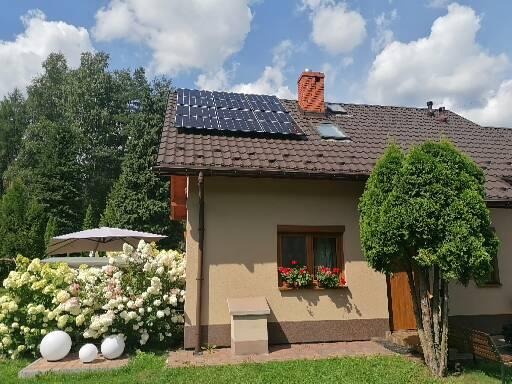 Dom jednorodzinny z brązowym dachem, na którym zamontowane są panele słoneczne. W oknie widoczne czerwone kwiaty w skrzynkach, a obok wejścia rośnie duży krzew.