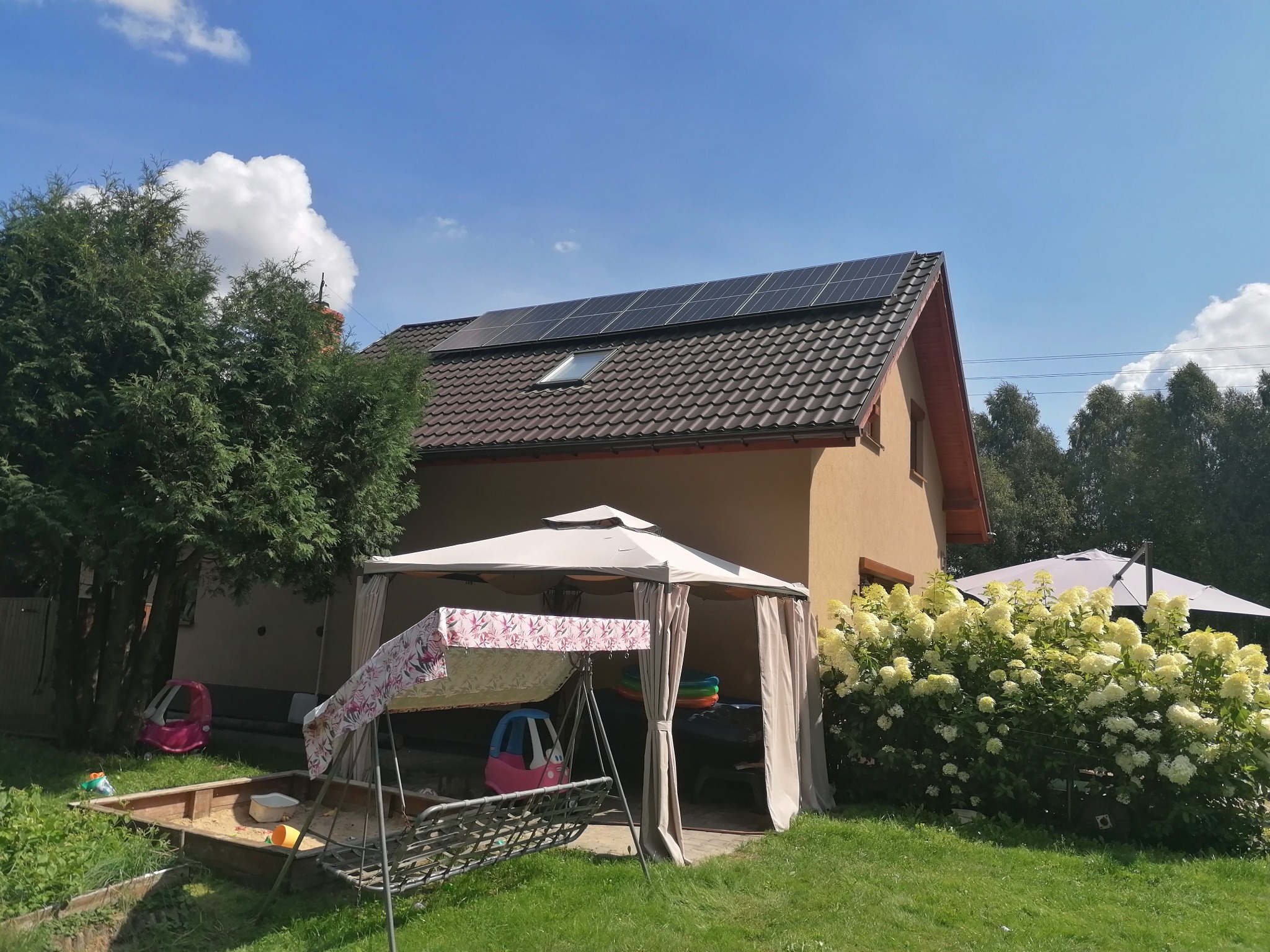 Dom jednorodzinny z brązowym dachem i panelami słonecznymi, widok z ogrodu z huśtawką i piaskownicą na pierwszym planie.