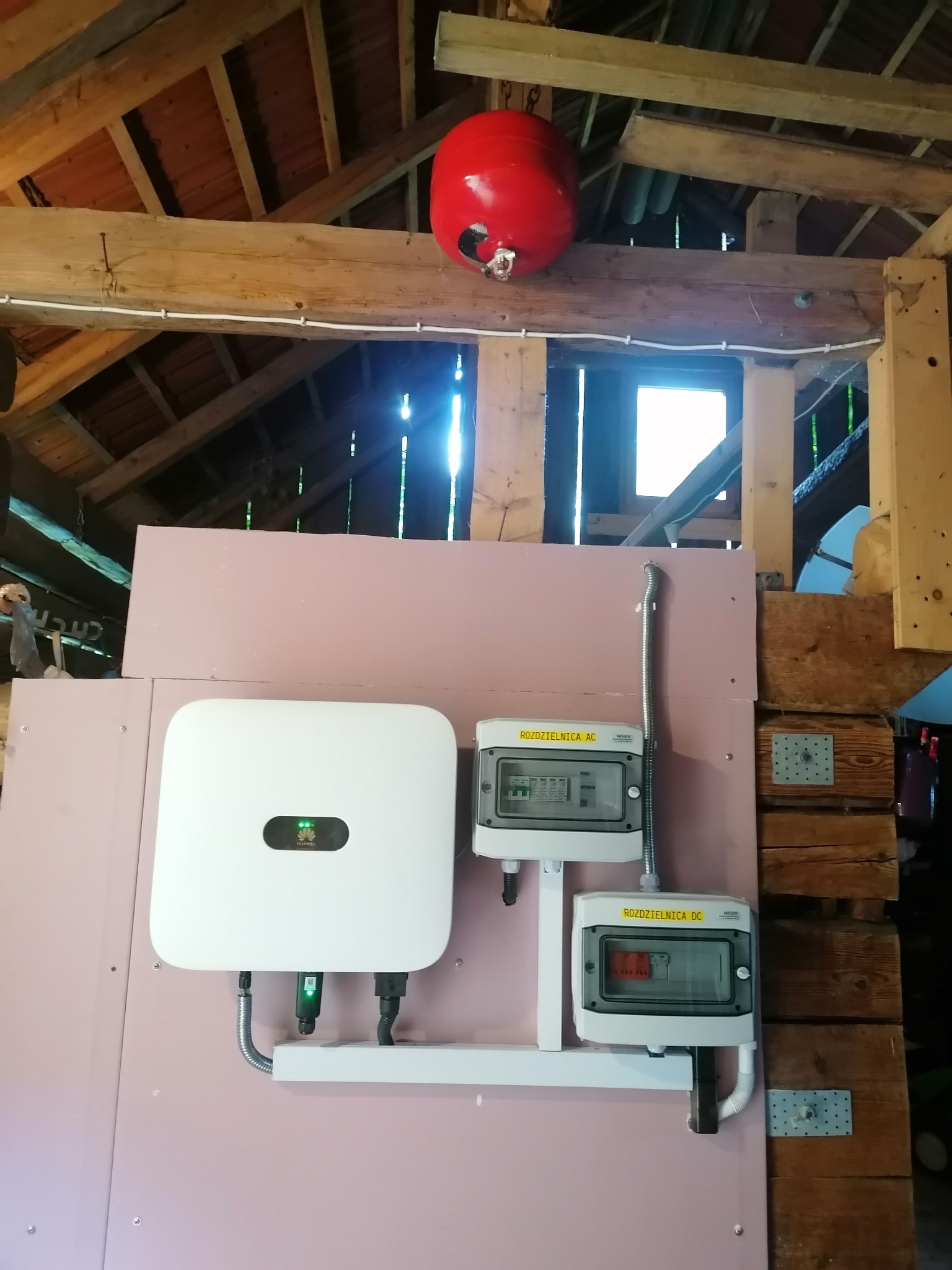 Biały falownik Huawei i dwie skrzynki rozdzielcze AC/DC zamontowane na różowej płycie OSB w surowym wnętrzu poddasza z drewnianą konstrukcją dachu i czerwoną kulą hydroforu.