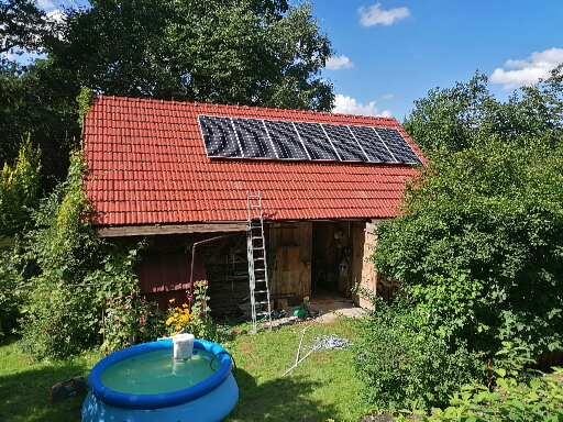 Panele słoneczne na czerwonym, spadzistym dachu małego budynku gospodarczego z drabina opartą o ścianę, na pierwszym planie mały basen.