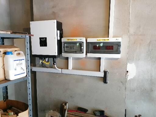 Biała skrzynka inwertera z podłączonymi kablami, zamontowana obok dwóch skrzynek z bezpiecznikami na szarej ścianie w pomieszczeniu gospodarczym.