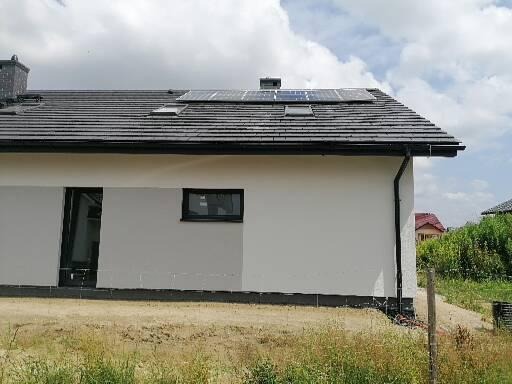 Panele słoneczne na dachu domu jednorodzinnego z szarą dachówką, widok z poziomu gruntu w pochmurny dzień.