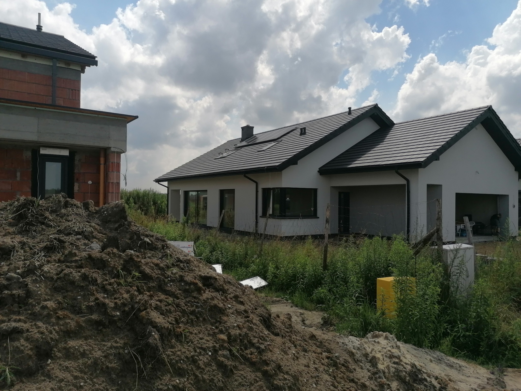 Dwa domy w stanie surowym, jeden z zamontowanymi panelami słonecznymi na dachu, na pierwszym planie hałda ziemi.