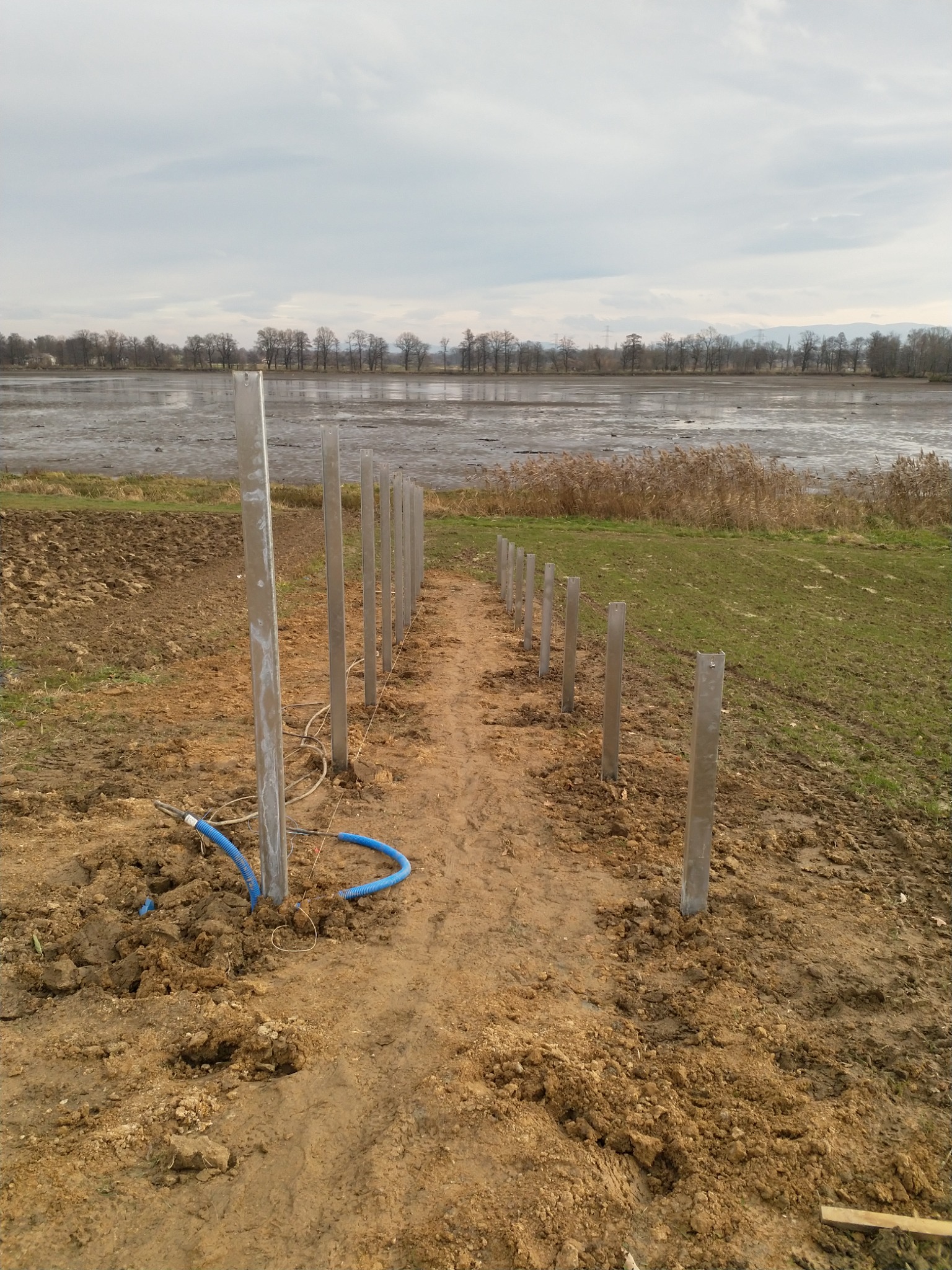 Przygotowanie terenu pod instalację, widoczne metalowe słupki w ziemi, niebieskie kable, w tle zbiornik wodny i linia drzew na horyzoncie, pochmurne niebo.