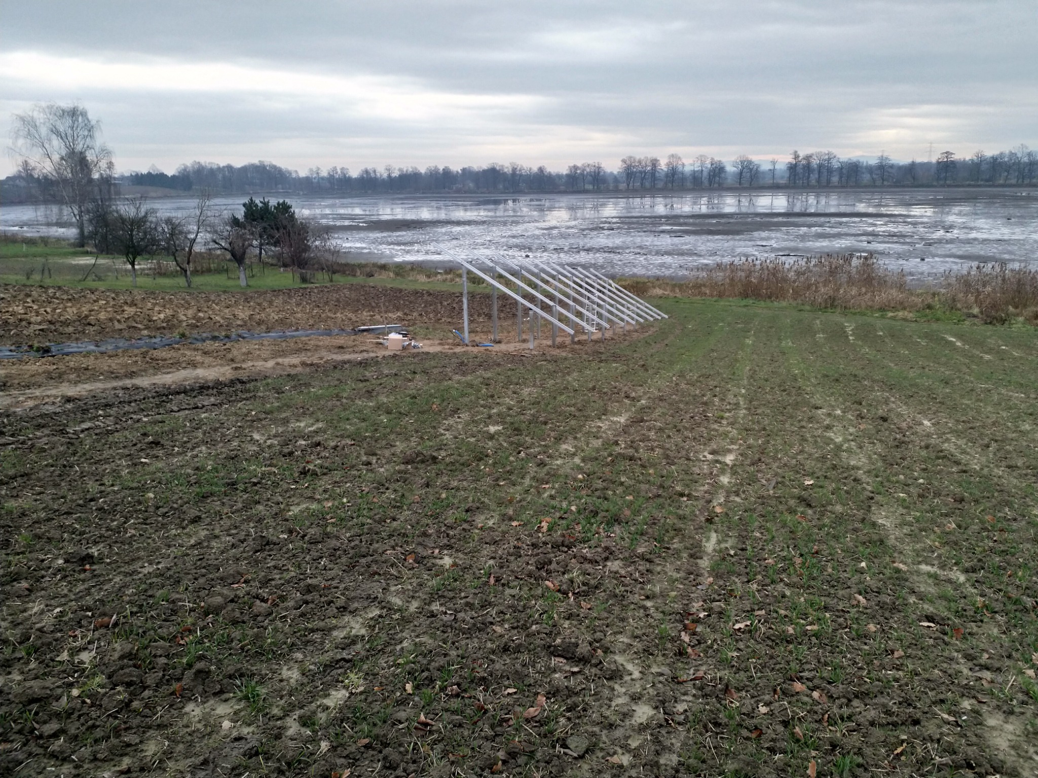 Stelaż konstrukcji pod panele słoneczne na polu uprawnym, w tle widoczne rozlewisko wodne i drzewa, pochmurny dzień.