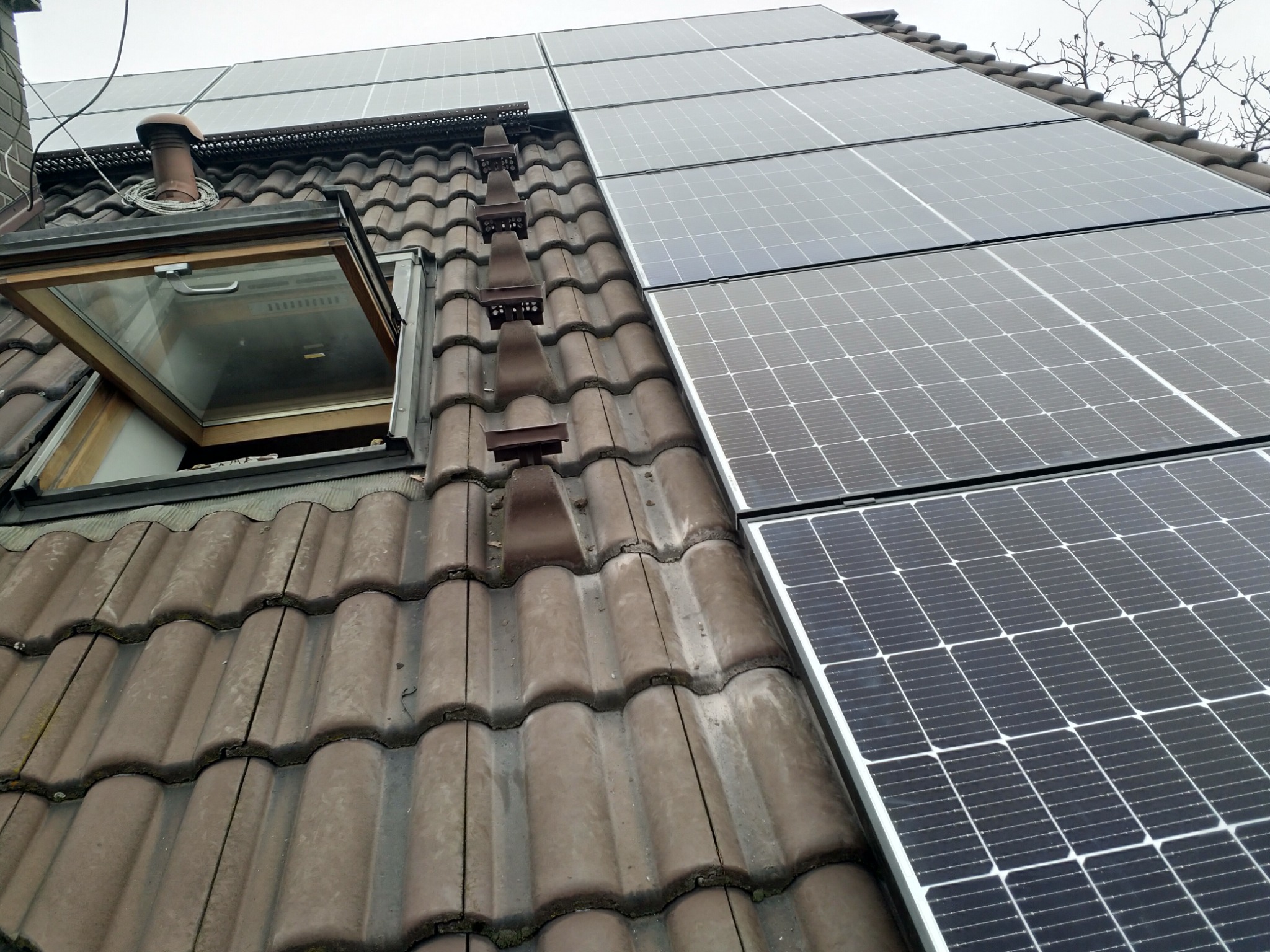 Panele słoneczne zamontowane na brązowym dachu z dachówki ceramicznej, widoczne okno dachowe i drabinka dachowa.