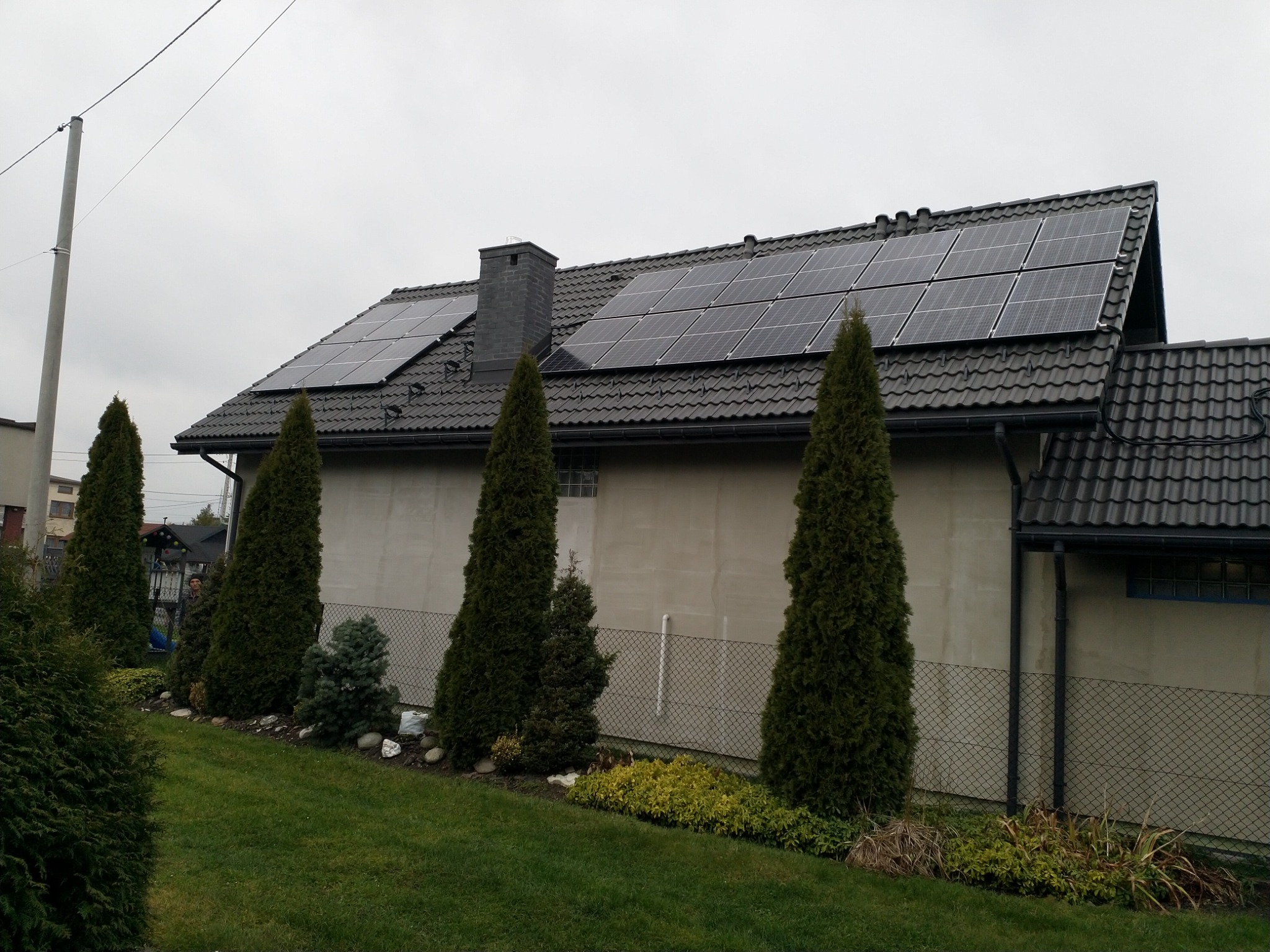 Dom jednorodzinny z ciemną dachówką i panelami słonecznymi na dachu, widok z ogrodu z tujami i trawnikiem.