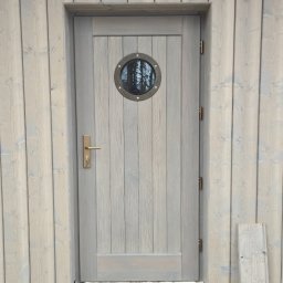 STOL-DACH MARCIN GRABOWSKI - Drewniane drzwi z okrągłym okienkiem w szarej, drewnianej ścianie. Widoczne zawiasy, klamka z kluczem i fragment deski leżącej obok. Styl marynistyczny.