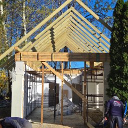 STOL-DACH MARCIN GRABOWSKI - Budowa altany w trakcie realizacji: drewniana konstrukcja dachu, ściany z bloczków, pracownicy z piłą łańcuchową. Widoczne drzewa w tle i błękitne niebo.