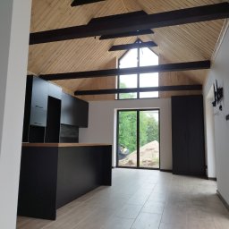 STOL-DACH MARCIN GRABOWSKI - Nowoczesne wnętrze z drewnianym stropem i czarną zabudową kuchenną. Duże okno z widokiem na zieleń. Minimalistyczny design, jasne płytki podłogowe.