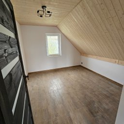 STOL-DACH MARCIN GRABOWSKI - Poddasze z drewnianą boazerią na suficie i podłogą z płytek imitujących drewno. Widoczne okno i czarne drzwi z białymi wstawkami. Nowoczesna lampa sufitowa.