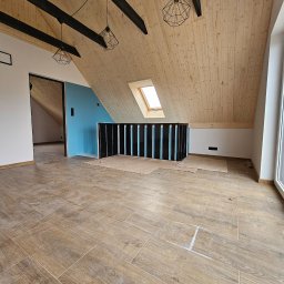 STOL-DACH MARCIN GRABOWSKI - Poddasze po remoncie: drewniana boazeria, czarne belki, okno dachowe, brązowe płytki podłogowe imitujące drewno. Widoczne ślady po pracach wykończeniowych.