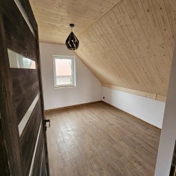 STOL-DACH MARCIN GRABOWSKI - Wykończone poddasze z drewnianą boazerią na skosach, brązowe panele podłogowe imitujące drewno, okno i nowoczesna lampa. Widok zza otwartych drzwi.