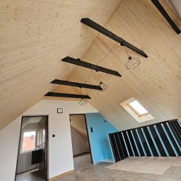 STOL-DACH MARCIN GRABOWSKI - Wykończone poddasze z drewnianą boazerią, czarnymi belkami i nowoczesnym oświetleniem. Widoczne okno dachowe i fragment łazienki. Podłoga wyłożona płytkami.