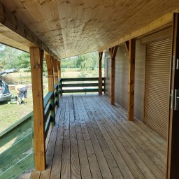 STOL-DACH MARCIN GRABOWSKI - Zadaszony taras drewniany z zieloną balustradą i widokiem na zewnątrz. Osoba kosi trawę w tle. Drewniane kolumny i rolety zewnętrzne.