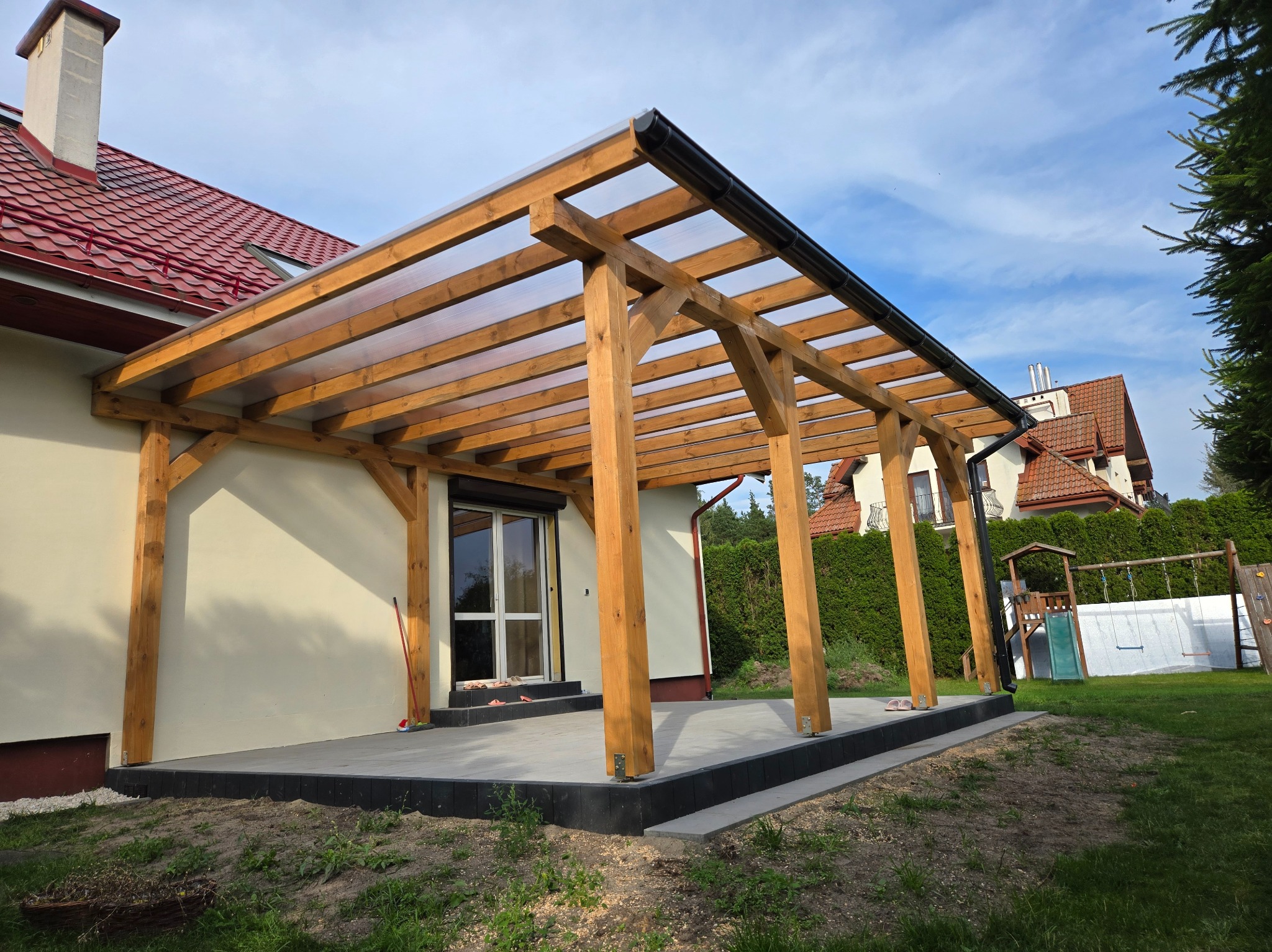 Drewniana pergola z przezroczystym zadaszeniem przylegająca do domu z czerwonym dachem. Widoczny fragment tarasu i plac zabaw w tle. Słoneczny dzień.
