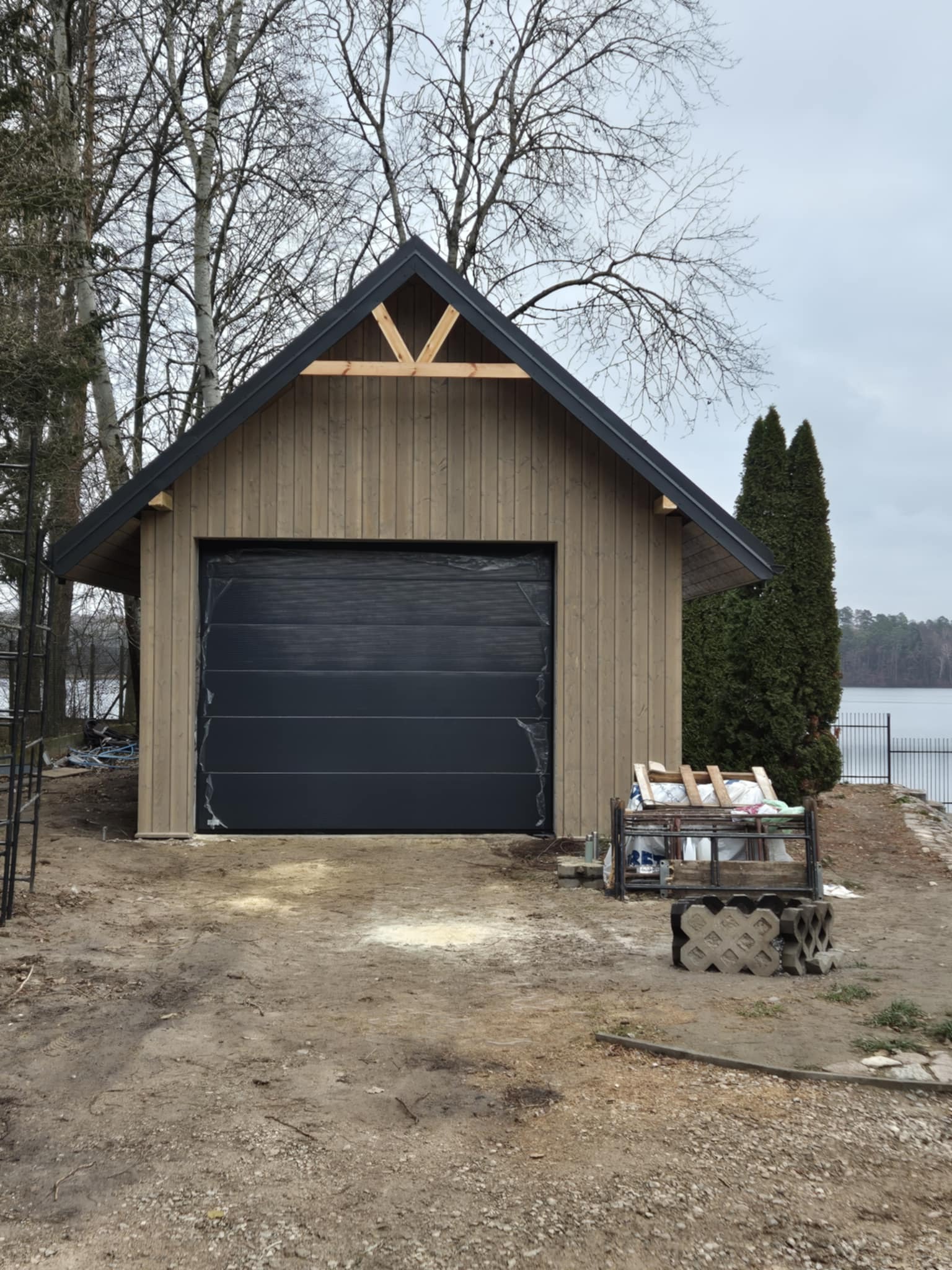 Nowa, drewniana altana z czarną bramą garażową na tle jeziora i drzew. Widoczne materiały budowlane. Świeżo postawiona konstrukcja.