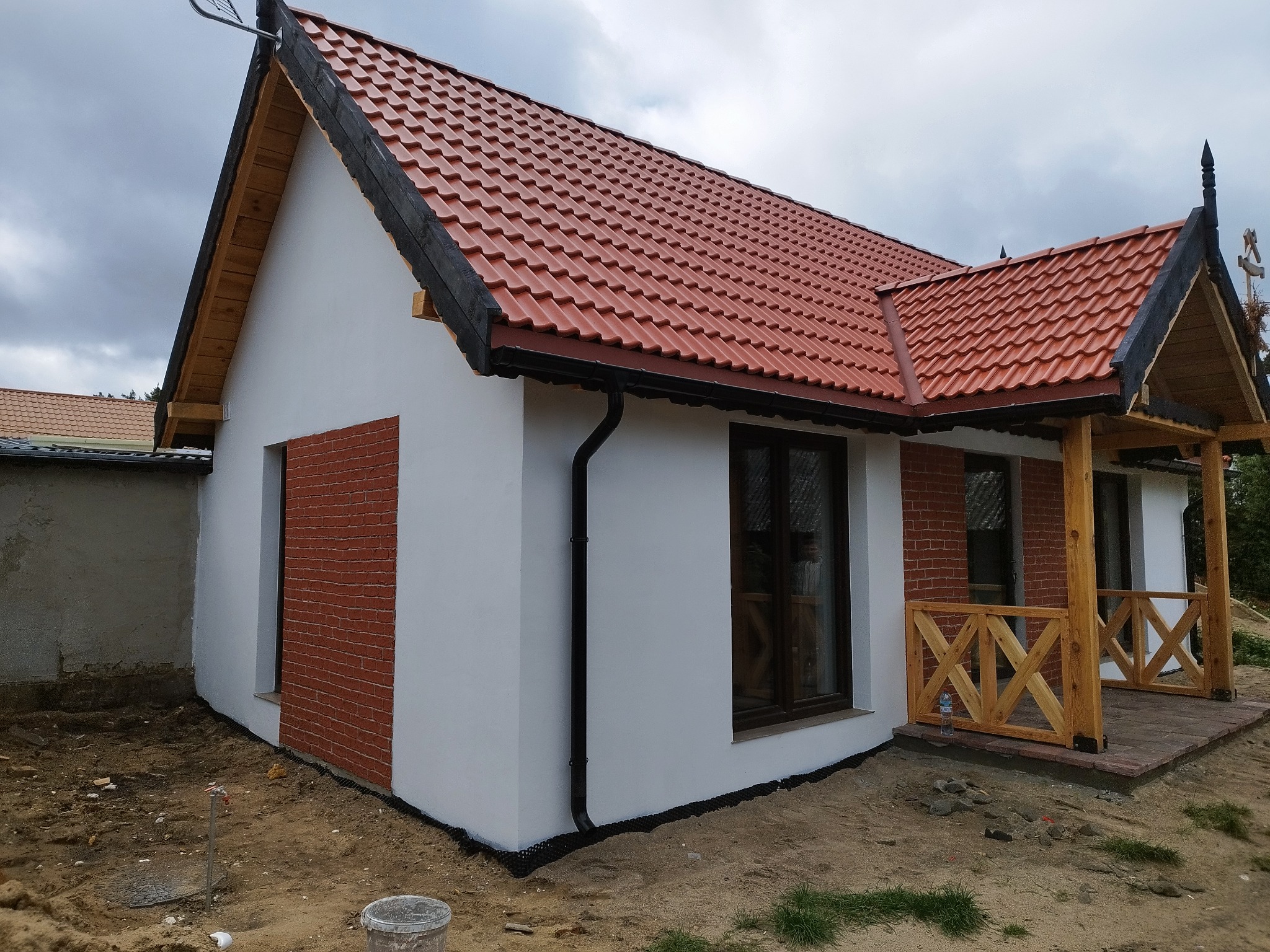 Nowoczesny dom jednorodzinny z czerwoną dachówką, białymi ścianami i elementami dekoracyjnymi z cegły. Widoczny drewniany taras i czarna rynna. Dom w trakcie wykończenia.