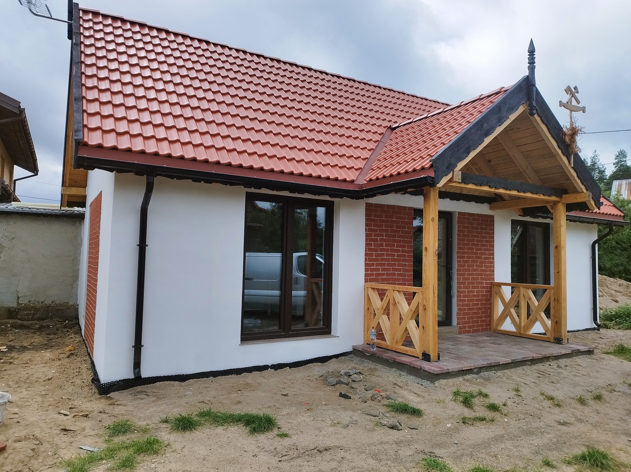 Nowy dom z czerwoną dachówką, białymi ścianami i drewnianą werandą z dekoracyjnym symbolem na dachu. Widoczny fragment elewacji z cegły.