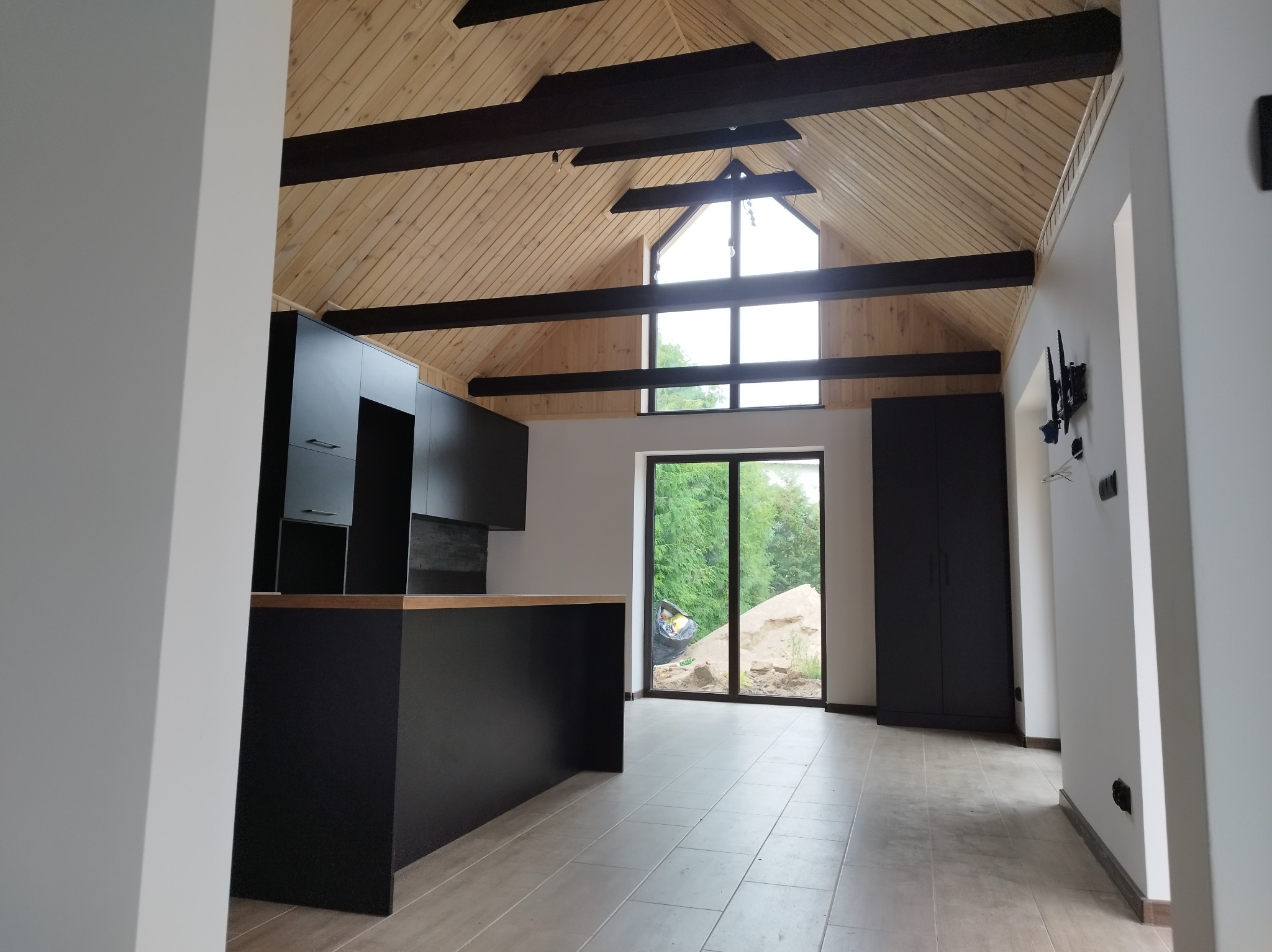 Nowoczesne wnętrze z drewnianym stropem i czarną zabudową kuchenną. Duże okno z widokiem na zieleń. Minimalistyczny design, jasne płytki podłogowe.