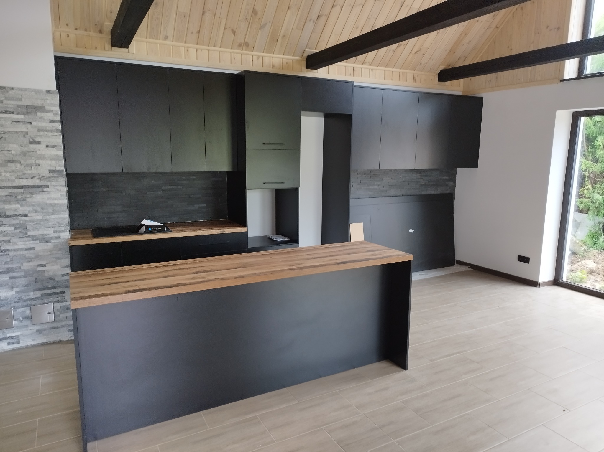 Nowoczesna kuchnia z czarnymi szafkami i drewnianym blatem wyspy, wykończona kamienną okładziną ściany i drewnianym sufitem z ciemnymi belkami.