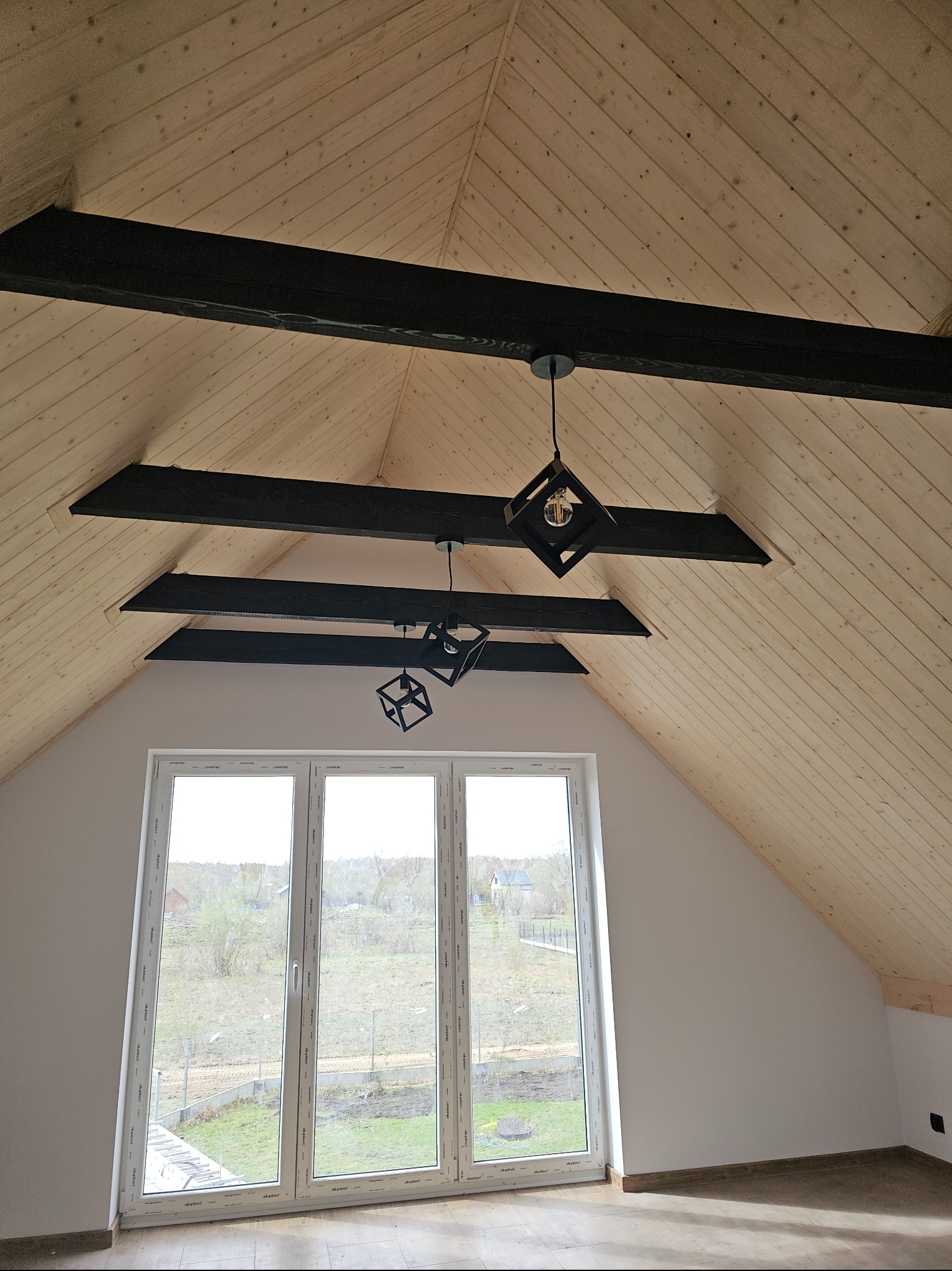 Poddasze z drewnianą boazerią, czarnymi belkami i nowoczesnymi lampami kostkami. Okno dachowe z widokiem na zewnątrz. Wykończenie wnętrza w jasnych barwach.