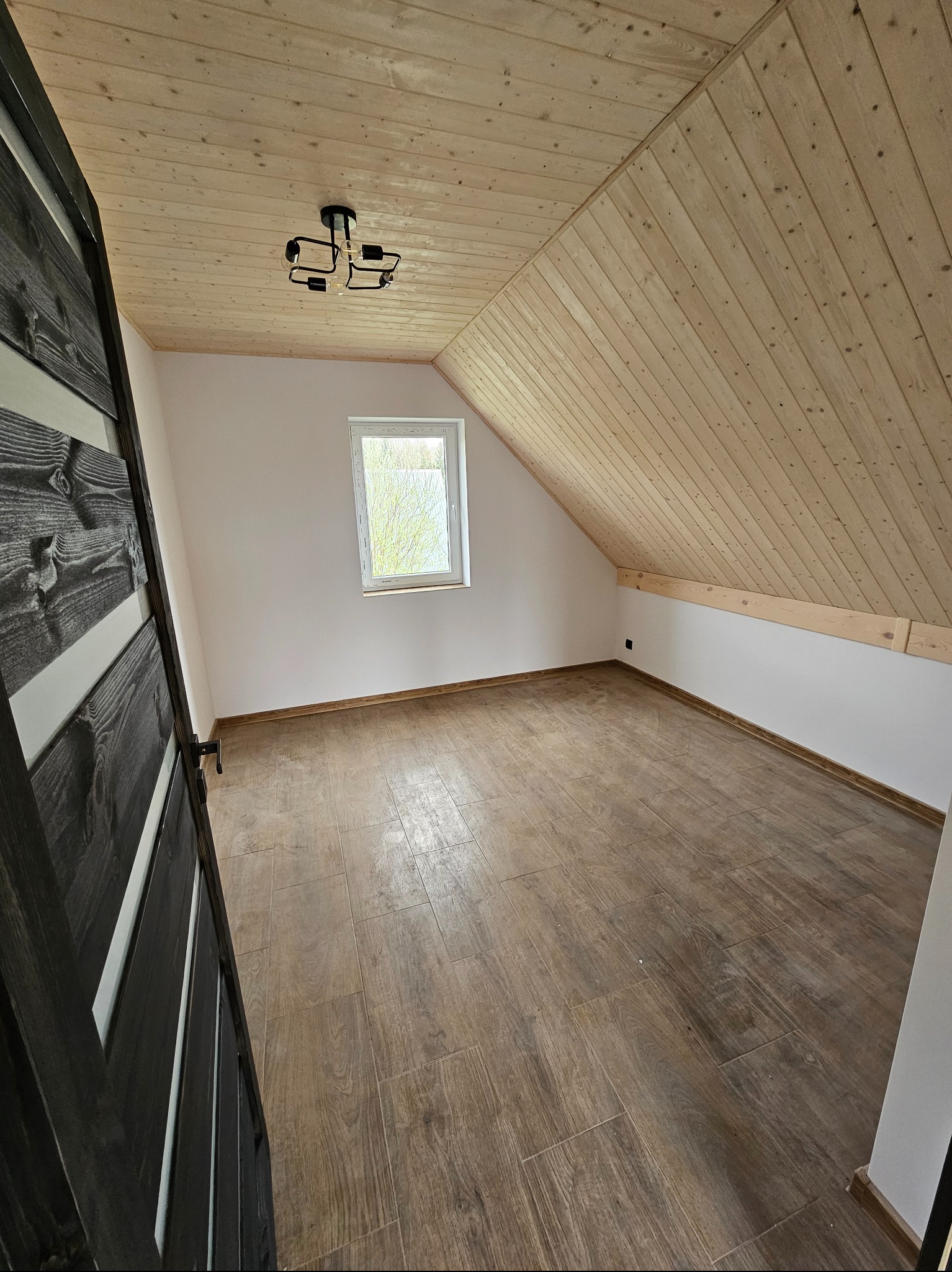 Poddasze z drewnianą boazerią na suficie i podłogą z płytek imitujących drewno. Widoczne okno i czarne drzwi z białymi wstawkami. Nowoczesna lampa sufitowa.