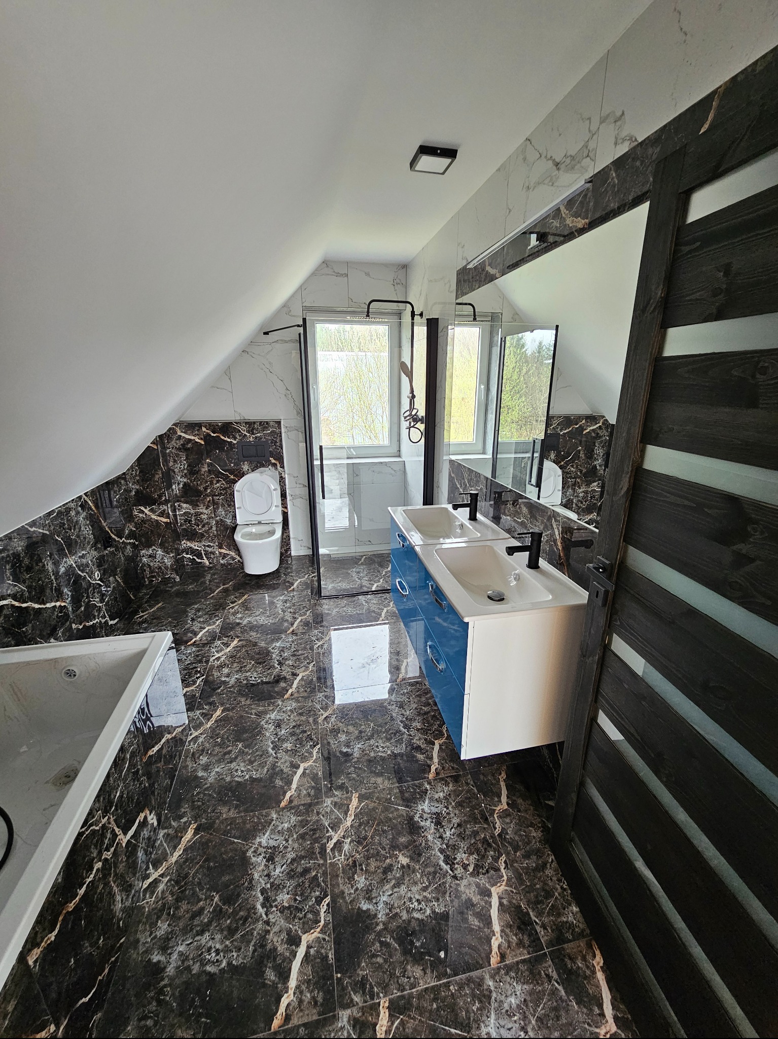 Wykończona łazienka na poddaszu w Pisz. Uwagę przykuwają marmurowe płytki w odcieniach bieli i brązu, kabina prysznicowa typu walk-in oraz szafka z dwiema umywalkami.