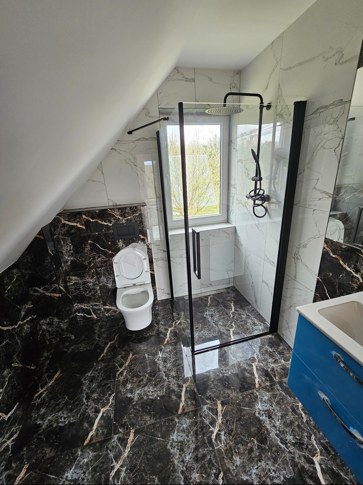 Wykończona łazienka na poddaszu z czarnymi marmurowymi płytkami, kabiną prysznicową typu walk-in z czarnymi ramami i białą ceramiką. Okno doświetla wnętrze.