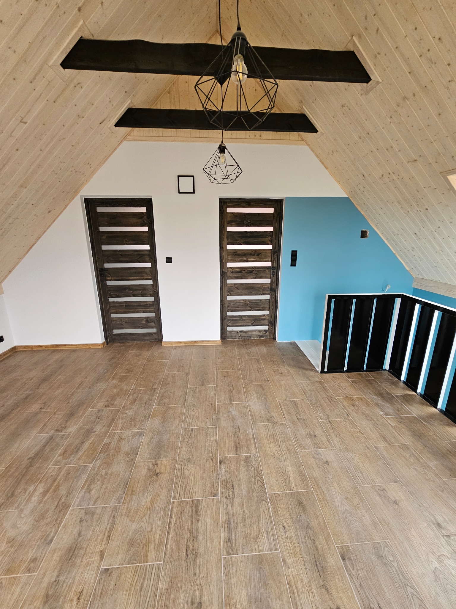 Wykończone poddasze z drewnianą boazerią, dwiema ciemnymi, nowoczesnymi drzwiami i drewnopodobną podłogą. Widoczna błękitna ściana i czarna balustrada.
