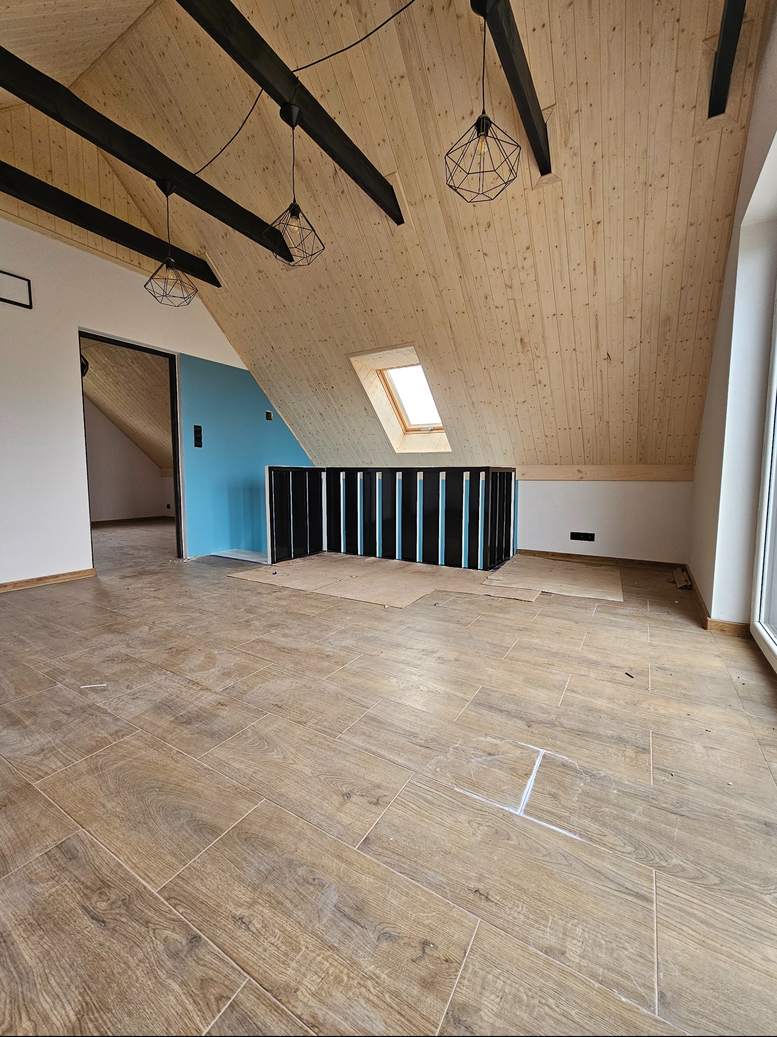 Poddasze po remoncie: drewniana boazeria, czarne belki, okno dachowe, brązowe płytki podłogowe imitujące drewno. Widoczne ślady po pracach wykończeniowych.