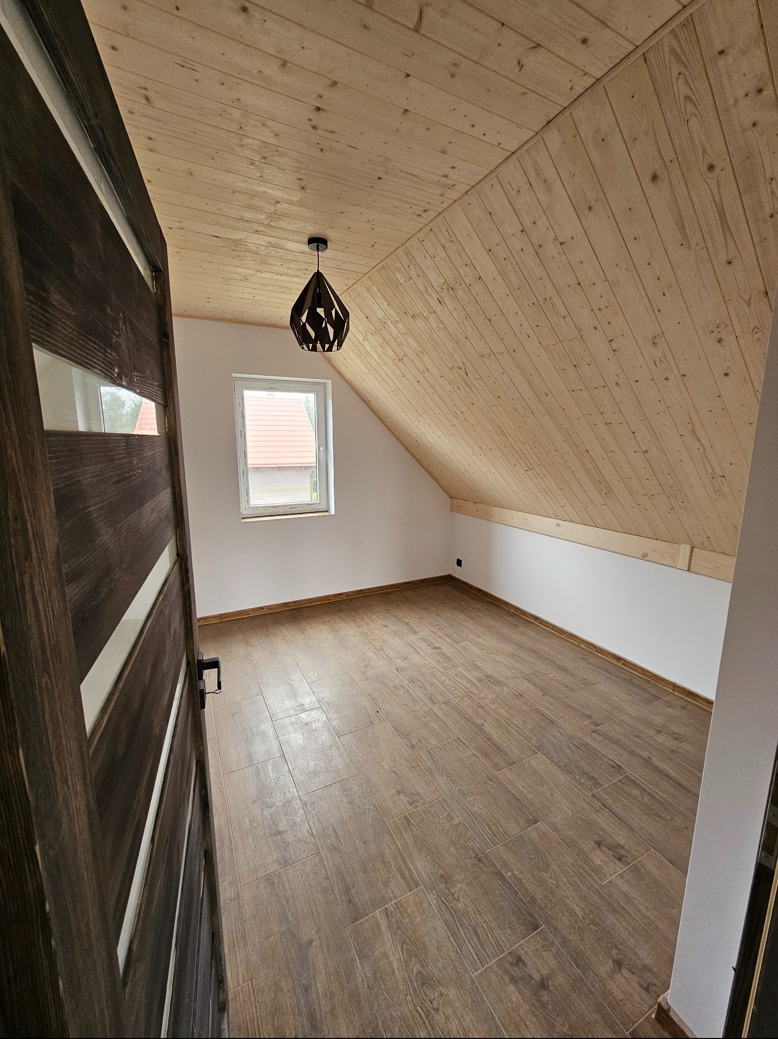 Wykończone poddasze z drewnianą boazerią na skosach, brązowe panele podłogowe imitujące drewno, okno i nowoczesna lampa. Widok zza otwartych drzwi.