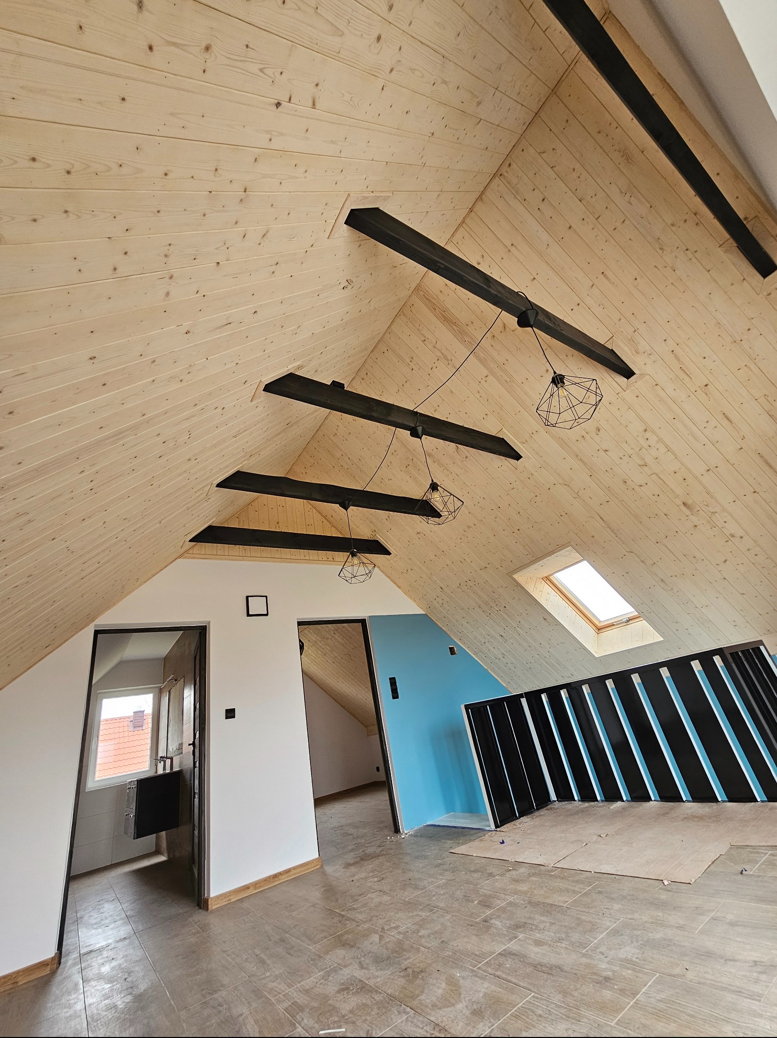Wykończone poddasze z drewnianą boazerią, czarnymi belkami i nowoczesnym oświetleniem. Widoczne okno dachowe i fragment łazienki. Podłoga wyłożona płytkami.