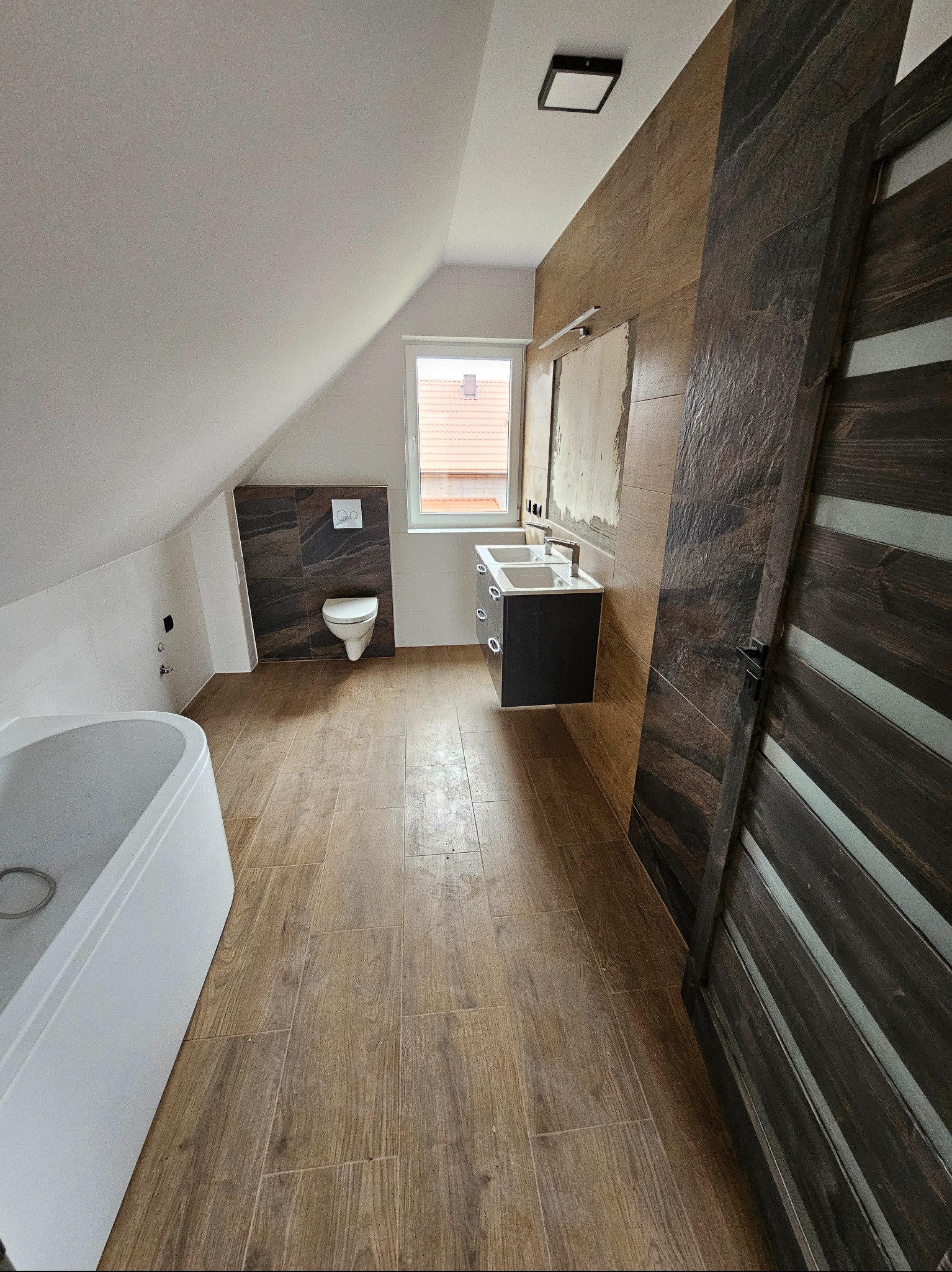 Ukończona łazienka na poddaszu z drewnopodobnymi płytkami, białą wanną i umywalką z szafką. Widoczne okno dachowe i częściowo wykończona ściana.