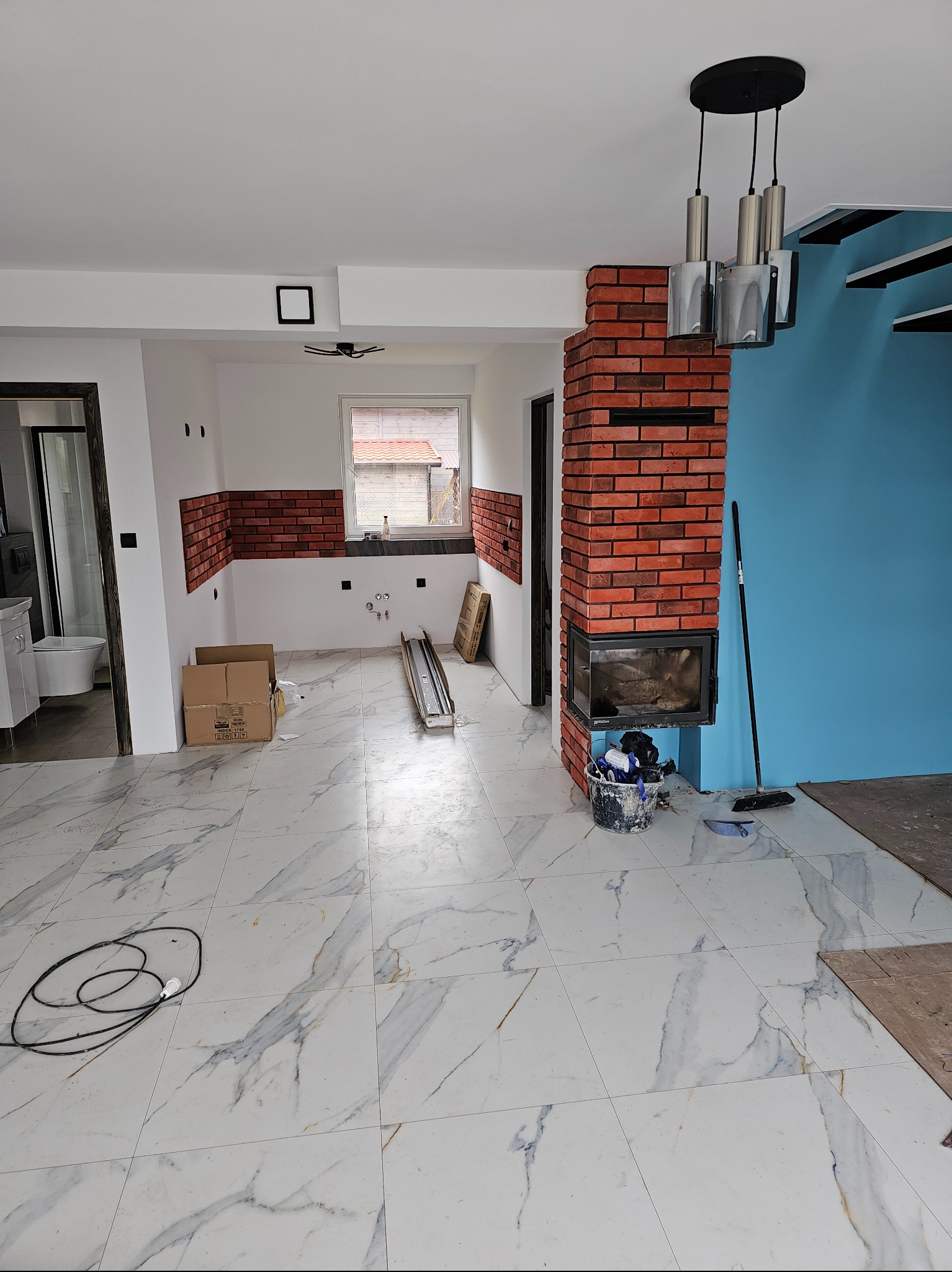 Wnętrze w trakcie remontu: marmurowe płytki podłogowe, fragment ściany z cegły, niebieska ściana, widoczne narzędzia i materiały budowlane. W tle łazienka.