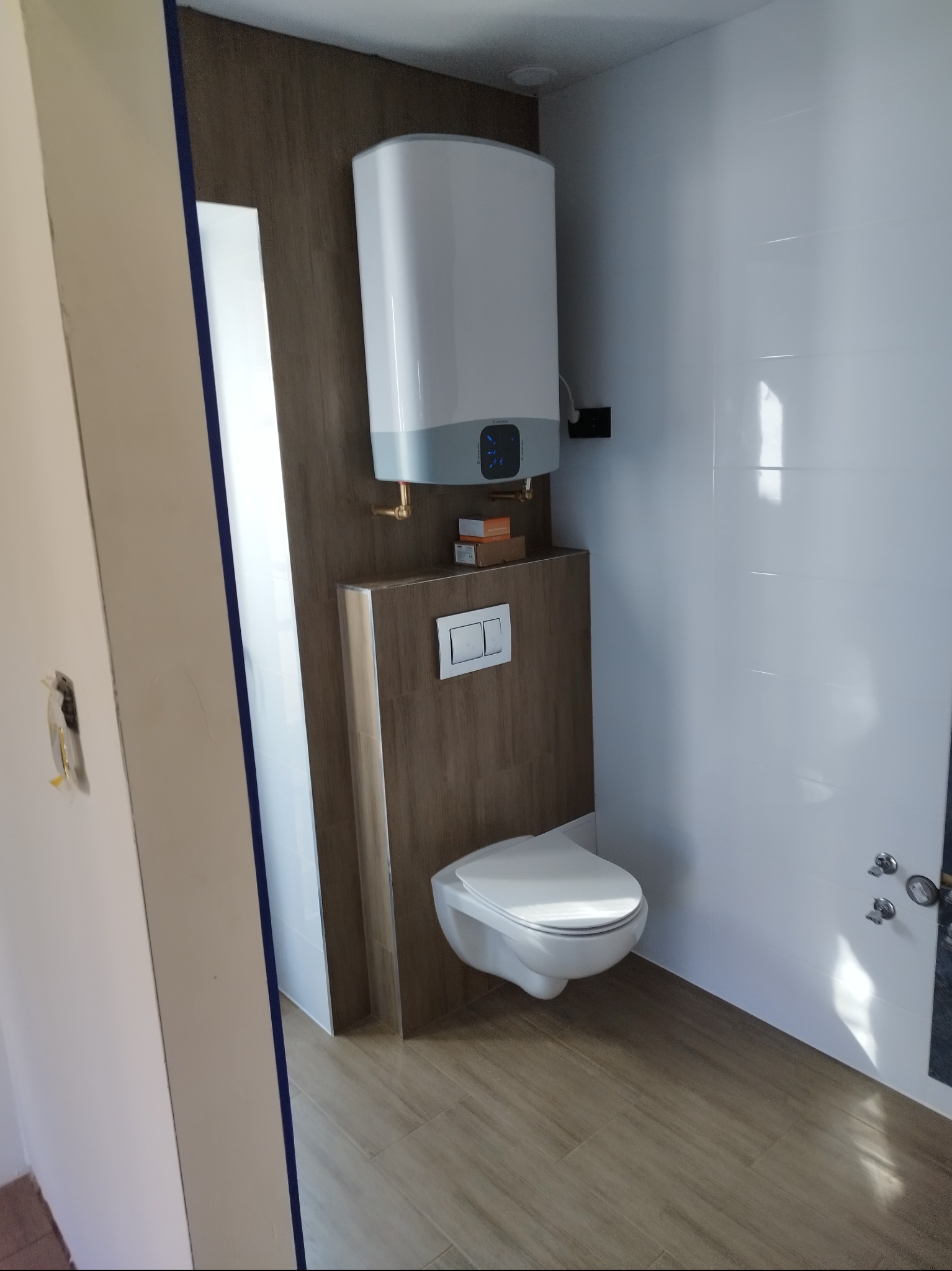 Nowoczesna łazienka po remoncie: podwieszana toaleta, bojler, brązowe panele ścienne i białe płytki. Widoczne detale wykończenia i armatura.