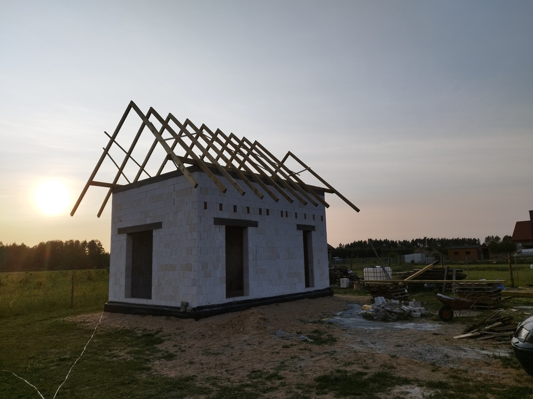 Surowy stan domu z białych bloczków, z widoczną konstrukcją dachu w trakcie budowy, na tle zachodzącego słońca i zieleni. Widoczne materiały budowlane i narzędzia.