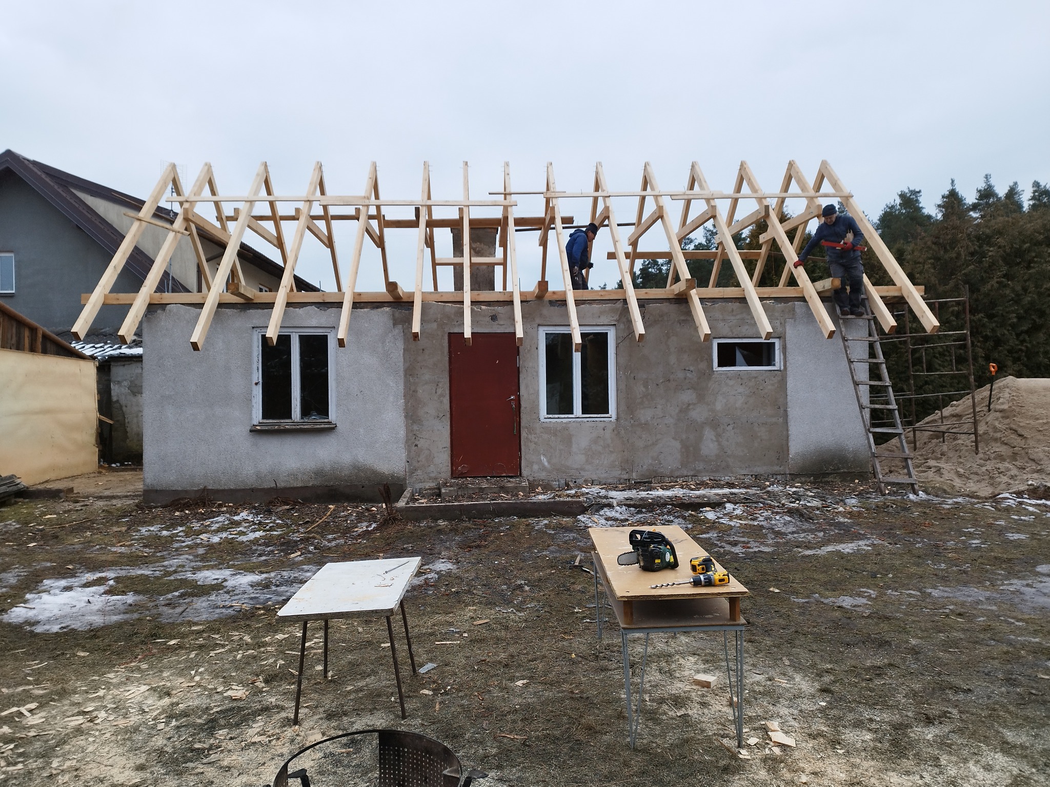 Budowa dachu: Dwóch pracowników montuje drewnianą konstrukcję więźby dachowej na murowanym budynku. Na pierwszym planie stoły z narzędziami.