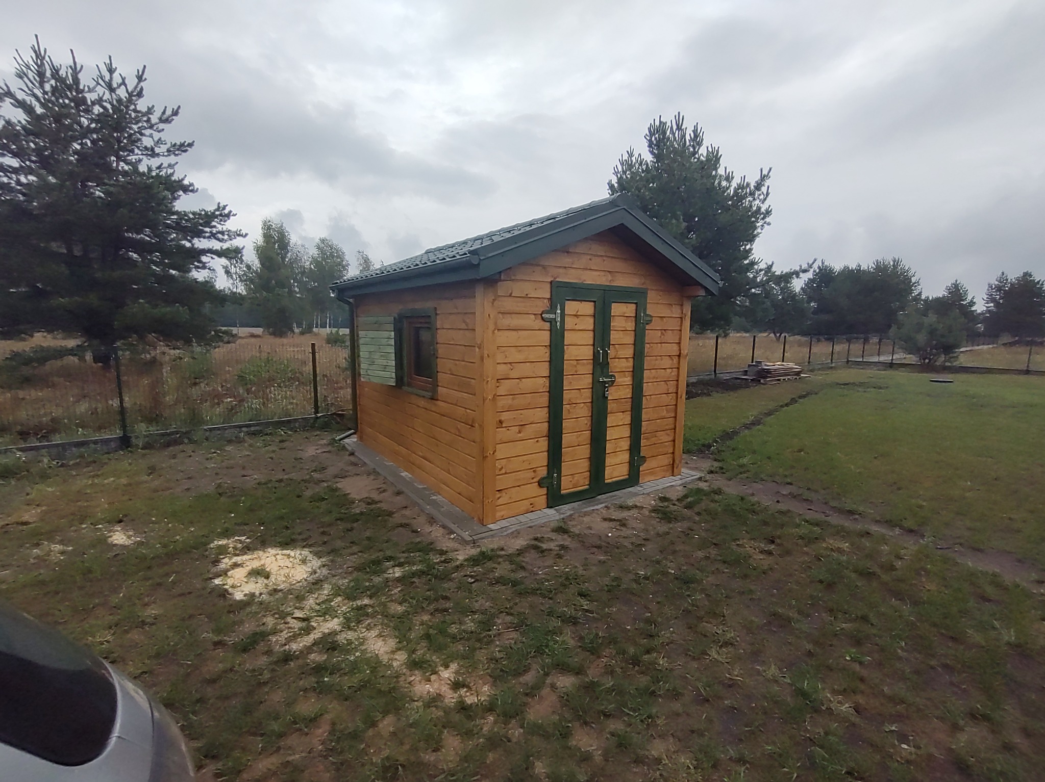 Drewniana altana ogrodowa z zielonymi akcentami, widoczna z boku na tle pochmurnego nieba i drzew. Świeże ślady montażu na ziemi.