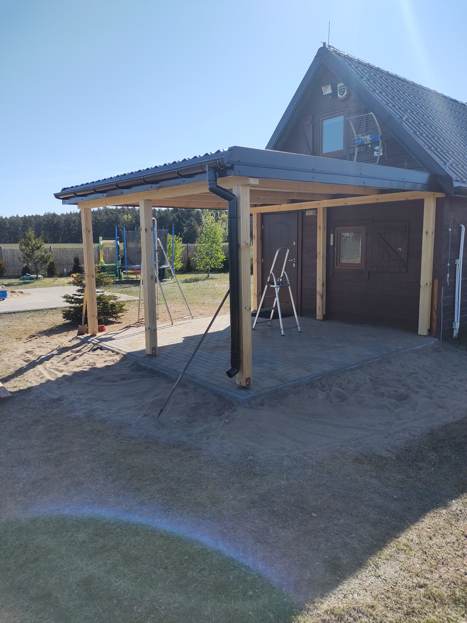 Drewniana konstrukcja zadaszenia tarasu przy brązowym domu z szarym dachem. Widoczne drabiny, teren przygotowany pod dalsze prace wykończeniowe.