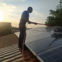 Mężczyzna w niebieskiej koszulce czyści panele słoneczne na dachu domu za pomocą szczotki na długim kiju, widoczny budynek w tle i drzewo po prawej, ujęcie z dołu pod światło.