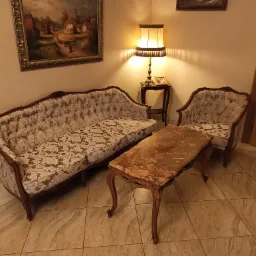 Antyczny komplet wypoczynkowy: sofa i fotel z kremowo-złotym wzorem kwiatowym, stolik kawowy z marmurowym blatem, oświetlony ciepłym światłem lampki nocnej, obraz w złotej ramie na ścianie.