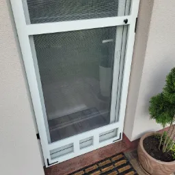 Białe drzwi moskitierowe z drobną siatką, czarnymi detalami w rogu i okrągłą gałką, widoczne z góry, z wycieraczką i doniczką obok.