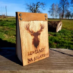 Drewniana książka z wypalonym wizerunkiem głowy jelenia i napisem 'LOVELASY NA SZLAKU', z brązową skórzaną wstawką i ozdobnymi metalowymi ćwiekami, ustawiona na drewnianym blacie na tle zielonej...